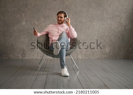 Handsome man listening to music while sitting in comfortable armchair against gray wall