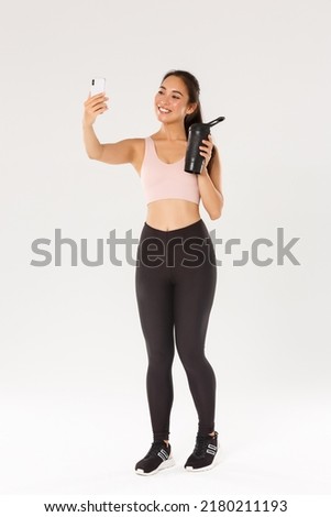 Full length of smiling slim and healthy asian girl taking selfie fitness training session, female blogger and athelte showing water bottle on camera of mobile phone, standing over white background