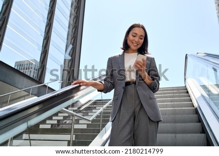 Smiling young Asian business woman wearing suit standing on urban escalator using applications on cell phone, reading news on smartphone, fast connection, checking mobile apps outdoors. Royalty-Free Stock Photo #2180214799