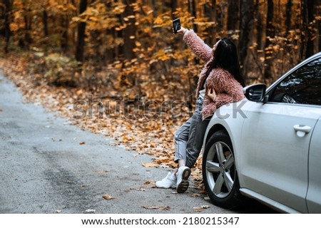 Confident brunette woman standing near car and doing selfie on cell phone. Fall autumn road weekend