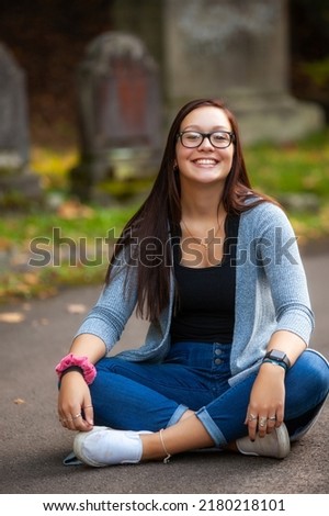 portrait of a pretty teenaged girl with dark hair and glasses seated in a cemetery Royalty-Free Stock Photo #2180218101