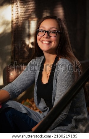 portrait of a pretty teenaged girl with dark hair and glasses seated on steps in the shadows in a park with beams of sunlight shining through Royalty-Free Stock Photo #2180219185