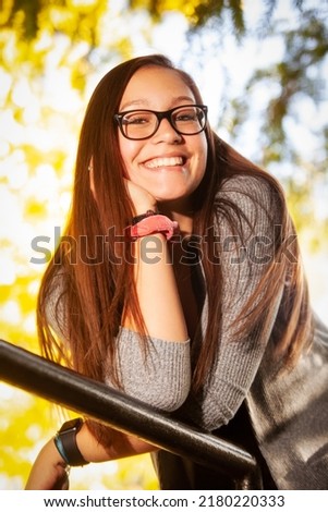 portrait of a pretty teenaged girl with dark hair and glasses in a park leaning o an iron rail in autumn Royalty-Free Stock Photo #2180220333