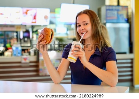 Happy positive smiling teen teenager girl, young woman is holding big juicy fat burger, hamburger and glass of soda sitting at table on food court. Fast food restaurant, junk unhealthy meal concept. Royalty-Free Stock Photo #2180224989