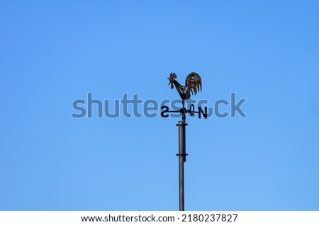 Weather vane or weathercock with wind direction indicator in the form of a compass rose on a roof against a blue sky. Royalty-Free Stock Photo #2180237827
