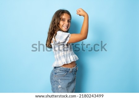 Smiling strong little brunette kid girl 10-11 years old showing biceps, muscles isolated on blue background children studio portrait. Childhood lifestyle concept. Mock up copy space. Royalty-Free Stock Photo #2180243499