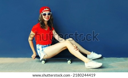 Modern happy smiling young woman sitting on skateboard wearing baseball cap, shorts on city street on blank copy space blue wall background