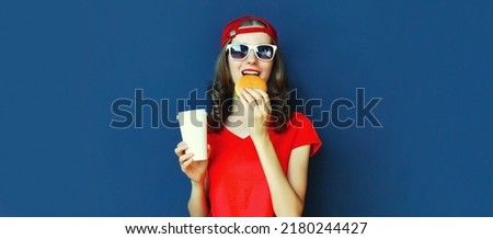 Portrait of happy young woman with fast food, burger and cup of juice wearing red t-shirt, sunglasses, baseball cap on blue background