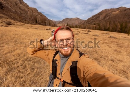 Concept Adventure Traveler with backpack. Hipster man in glasses and red hat takes selfie photo background autumn mountains Altai.
