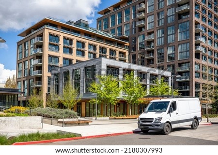 Commercial white compact cargo mini van for local delivery and small business standing on the urban city street with multilevel residential apartment buildings serving neighborhood Royalty-Free Stock Photo #2180307993