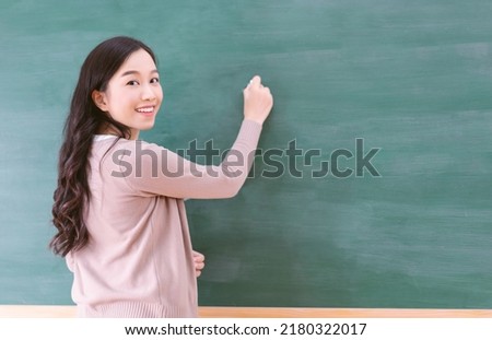 Portrait of smiling Asian teacher woman writing on chalkboard, teach english and math to students in classroom. Female teacher writing on blackboard wall and looking at camera. Back to school concept Royalty-Free Stock Photo #2180322017