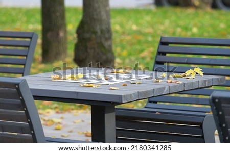 A serene picture of an empty metallic chess table surrounded by grey structured wooden benches ready to play chess in an autumn park full of fallen yellow leaves. Weekend in the autumn park.
