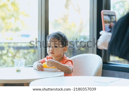Asian boy enjoying delicious orange cake in restaurant with a smartphone secretly taking pictures