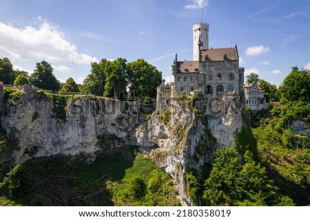 Aerial view of lichtenstein castle (Schloss) on forested rock cliff in Swabian Alps in summer. Seasonal panorama of romantic fairytale palace in Gothic revival style over sky. European famous landmark Royalty-Free Stock Photo #2180358019