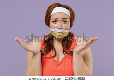 Young confused sad chubby overweight plus size big fat fit woman in red top gagged with measuring tape warm up training isolated on purple color background gym home Workout motivation sport concept. Royalty-Free Stock Photo #2180414825