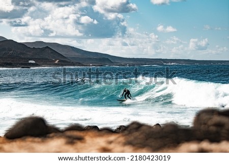 Wild rocky coastline of surf spot La Santa Lanzarote, Canary Islands, Spain. Surfer riding a big wave in rocky bay, volcano mountain in background.