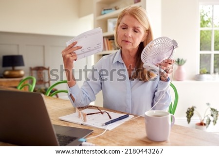 Menopausal Mature Woman Having Hot Flush At Home Cooling Herself With Fan Connected To Laptop Royalty-Free Stock Photo #2180443267