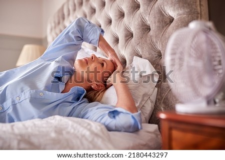 Menopausal Mature Woman Suffering With Insomnia In Bed At Home Using Electric Fan Royalty-Free Stock Photo #2180443297