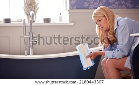 Menopausal Mature Woman Suffering With Incontinence Holding Pad Sitting On Toilet At Home Royalty-Free Stock Photo #2180443307
