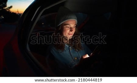 Side night cinematic view of young beautiful woman sit in taxi car back seat near window on road trip, hold, check on mobile phone, use smartphone, scroll social media news, read messages and smile Royalty-Free Stock Photo #2180444819