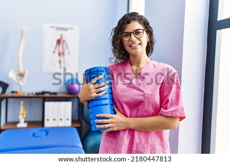 Young latin woman wearing physiotherapist uniform holding foam roller at clinic Royalty-Free Stock Photo #2180447813