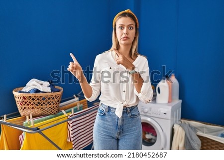 Young blonde woman at laundry room pointing aside worried and nervous with both hands, concerned and surprised expression 