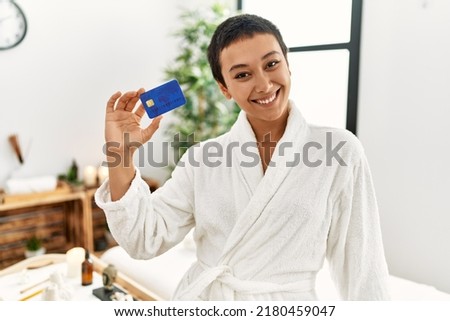Young hispanic woman wearing bathrobe holding credit card at beauty center