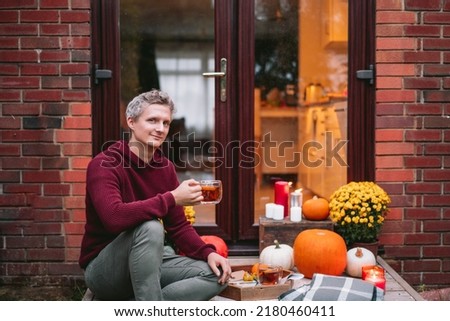 Portrait of young man with hot tea sitting on the porch of his home. Cozy ideas on how to spend time at home. Autumn tea time outdoors on house entrance decorated with pumpkins, flowers and candles.