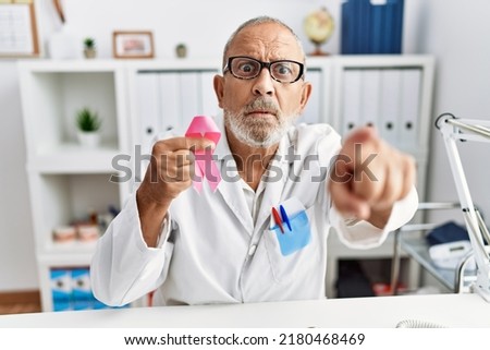 Mature doctor man holding pink cancer ribbon at the clinic pointing with finger to the camera and to you, confident gesture looking serious 