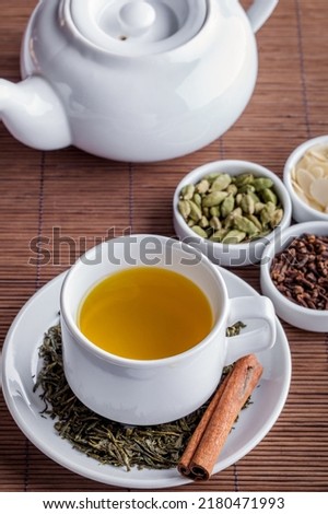 Green tea with sea buckthorn, cardamom, cloves, almond flakes and cinnamon. Tea in a white ceramic cup, on a white ceramic saucer. Green tea and a cinnamon stick are scattered on a saucer. Nearby lies