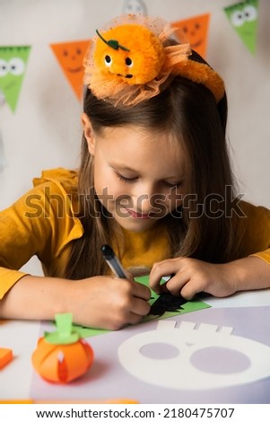 A little girl in a pumpkin costume draws a mask with a black felt-tip pen. Halloween party. Craft for kid