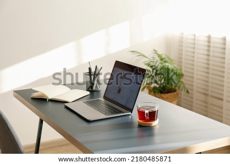 Home office concept. Designated work from home area near the window. Modern laptop with blurred screen and glass cup of tea on table. Close up, copy space, interior background. Royalty-Free Stock Photo #2180485871