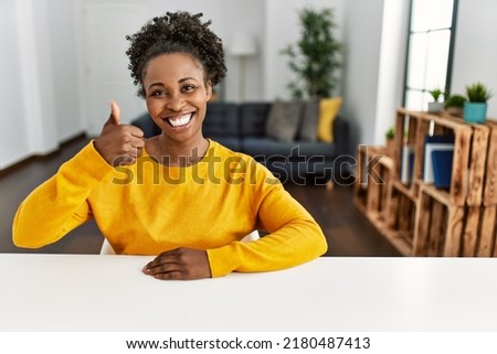 Young african american woman wearing casual clothes sitting on the table at home doing happy thumbs up gesture with hand. approving expression looking at the camera showing success. 