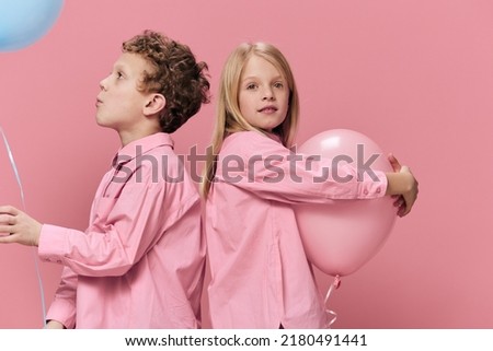 cute, happy kids in pink clothes on a pink background stand sideways to the camera and hug their big pink and blue balloons. Horizontal studio photo with empty space for advertising text insertion
