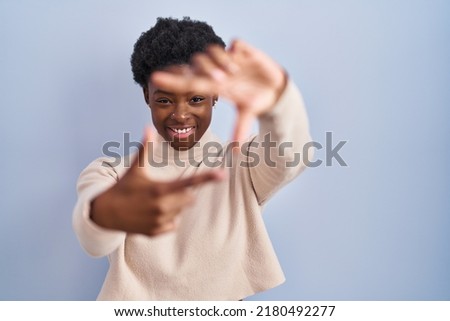 African american woman standing over blue background smiling making frame with hands and fingers with happy face. creativity and photography concept. 