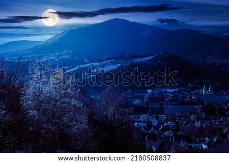 carpathian rural landscape in autumn at night. village in the valley at the foot of the mountain. beautiful countryside scenery in full moon light. trees in fall foliage on the grassy hills Royalty-Free Stock Photo #2180508837