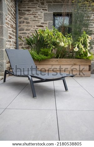Modern garden design and landscaping:Deck chair on a terrace paved with natural flagstones in contrast to a raised bed planted with bird of paradise and catalpa in wooden pot in front of a stone wall Royalty-Free Stock Photo #2180538805