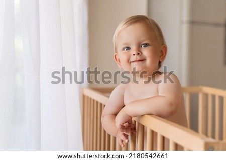 Indoor copy space portrait of adorable cute blue-eyed baby. Toddler standing in crib holding onto bumpers looking at his mom or dad with happy face and lovely smile. Royalty-Free Stock Photo #2180542661