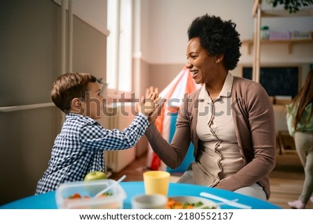 Happy little boy giving high-five to his African American teacher after eating lunch at kindergarten. Royalty-Free Stock Photo #2180552137