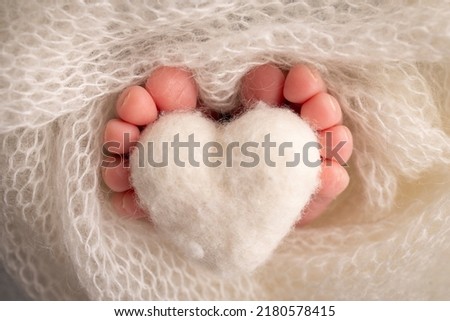 Knitted white heart in the legs of a baby. Soft feet of a new born in a white wool blanket. Close-up of toes, heels and feet of a newborn. Macro photography the tiny foot of a newborn baby. Royalty-Free Stock Photo #2180578415