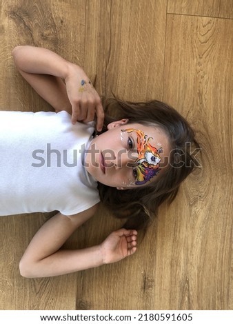 little girl is resting on a wooden floor and sad. hair of child is developing and she has face painting