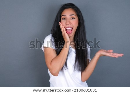 Positive glad Young hispanic  woman wearing white T-shirt over grey background says: wow how exciting it is, indicates something.  One hand on his head and pointing with other hand.