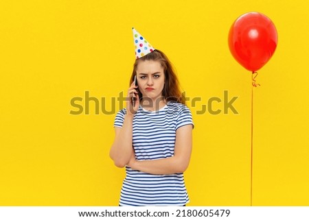 gloomy young woman is talking on a mobile phone, wears party cone and striped t shirt, feels embarrassed and offended has bad mood during celebration, standing over yellow background
