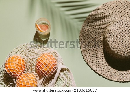 Summer flat lay with straw hat, glass of water and orange fruit in eco shopping bag. Green background with palm leaf shadow, sun and sunlight. Vacation, holiday, summer creative minimal concept Royalty-Free Stock Photo #2180615059