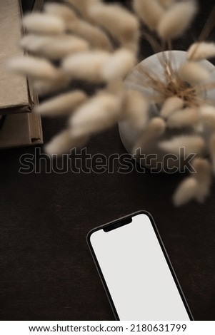 Aesthetic flatlay of blank screen mobile smart phone, books stack, bunny rabbit tail grass bouquet on deep brown background. Blog, social media, web, magazine copy space template. Flat lay, top view.