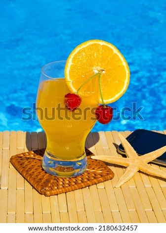 A refreshing cocktail, juice with a red cherry and an orange inside, next to a starfish and a mobile phone against the backdrop of a blue pool. On a summer sunny day.