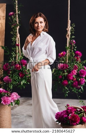pretty and happy woman in white cozy clothes near swing with peony flowers. florist services for room decor. comfortable home and summer clothes made of natural fabrics for women.