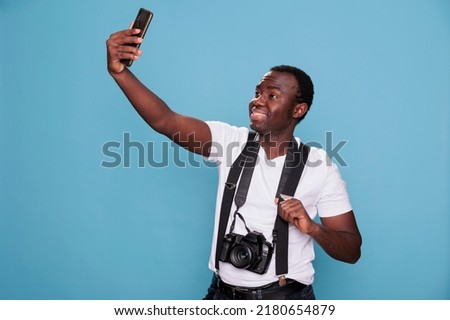 Cheerful photographer taking selfie photo with smartphone while smiling heartily on blue background. Confident and handsome photography enthusiast taking picture of himself while having DSLR camera.
