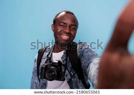 Happy photography enthusiast having professional photo device and taking selfie on blue background. Confident photographer with DSLR camera and backpack enjoying vacation.