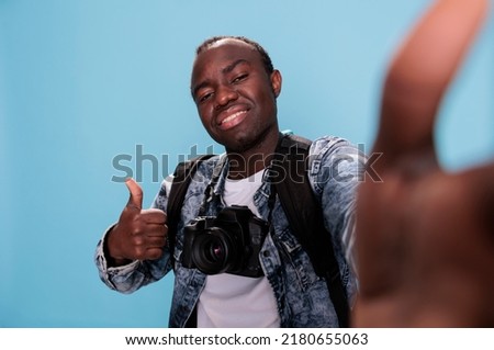Photography enthusiast giving approve hand gesture while taking picture of himself. Confident photographer showing okay finger symbol while taking selfie and having DSLR camera on blue background.